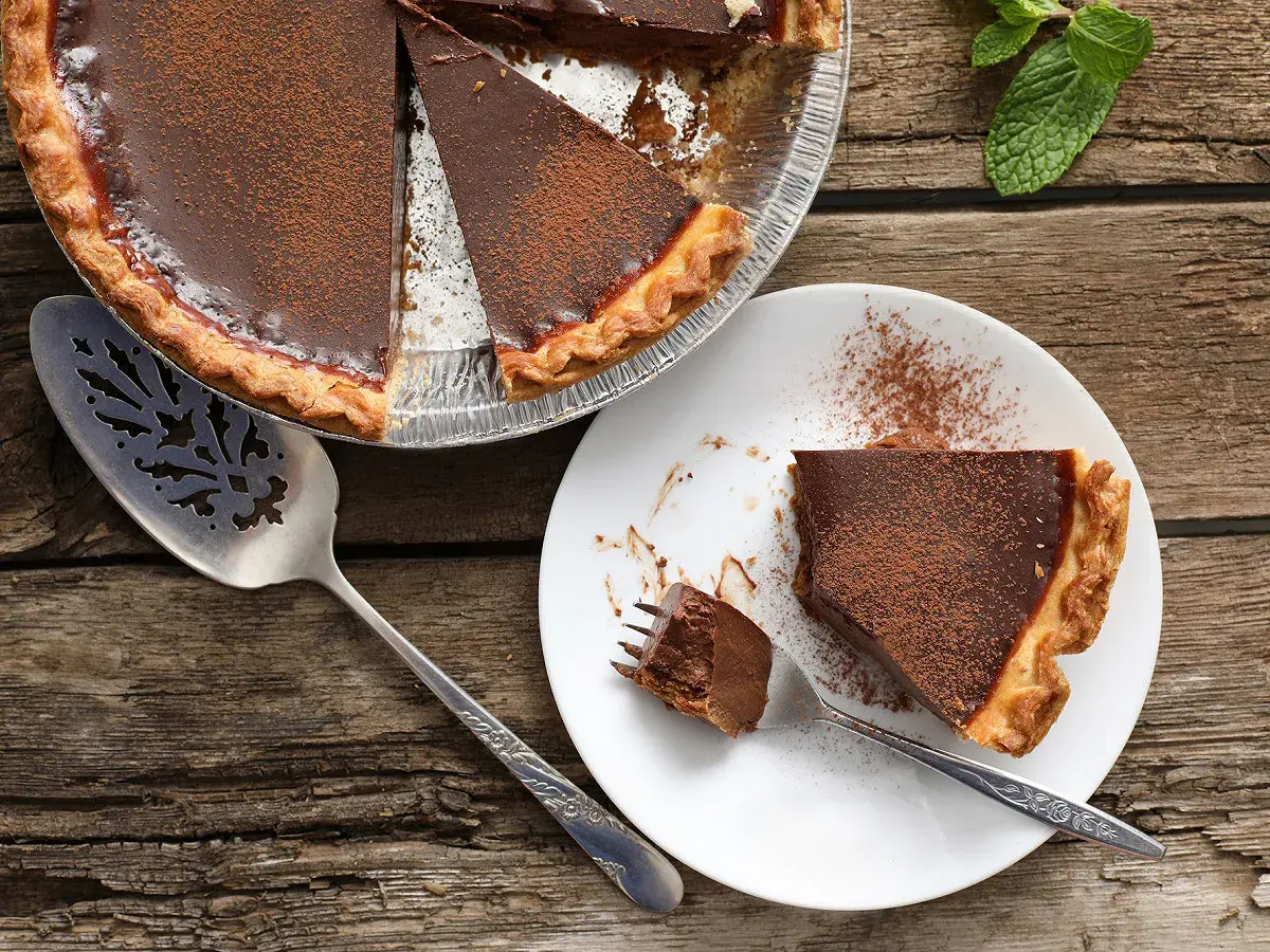 Une délicieuse tarte au chocolat, saupoudrée de cacao, est prête à être dégustée. Une part est servie sur une assiette blanche, avec une fourchette tenant un morceau.