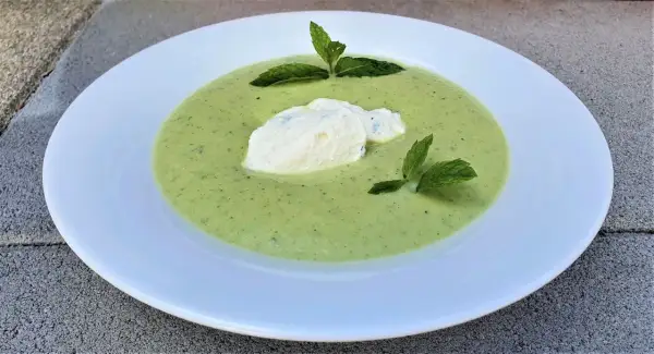 Velouté de courgette onctueux, garni d'une quenelle de fromage frais et de feuilles de menthe, servi dans une assiette blanche.