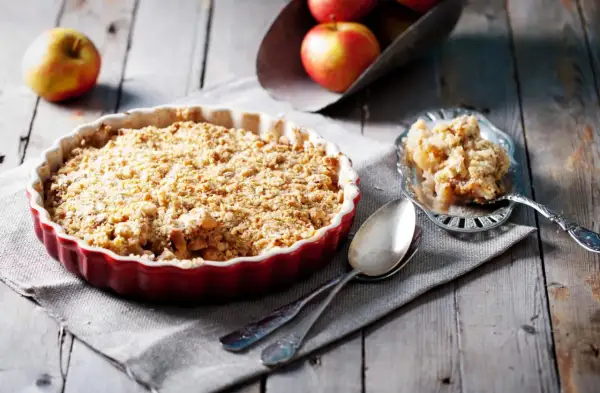 Un délicieux crumble aux pommes tout juste sorti du four, servi dans un plat rouge, avec une portion sur une cuillère ornée.